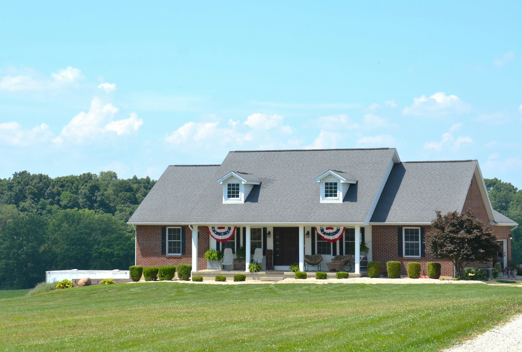 A brick house with a deep front porch, a flag, and a green lawn on a sunny day.